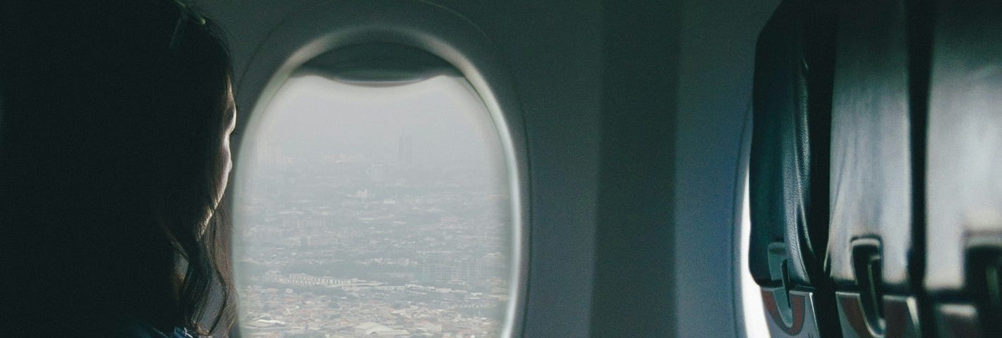 A woman, nearly silhouetted, young-ish, stares out the window of an airplane. Tones in photo are mostly greys and blues.