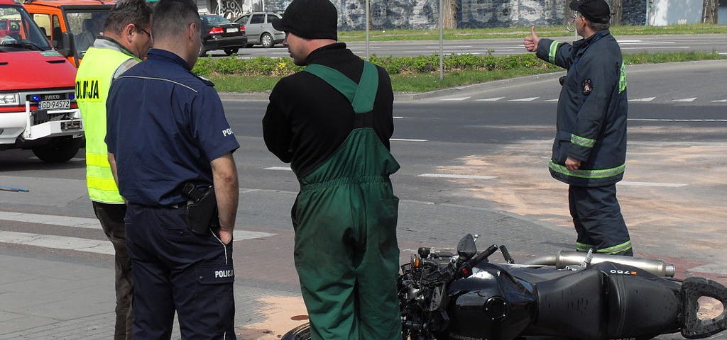 Black motorcycle seen laying on its side on a paved city road. The driver is standing and talking with some police officers. A fireman if directing the traffic around the scene.