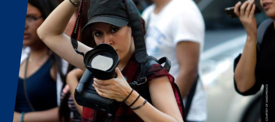 A woman photojournalist wearing a black baseball cap holds up a large camera and lifts her  other arm to hold up the camera’s strap that is around her neck. A message below the image states: For more than 30 years, USAID has supported independent media as part of its democracy promotion efforts. This includes bolstering the professionalism and safety of journalists.