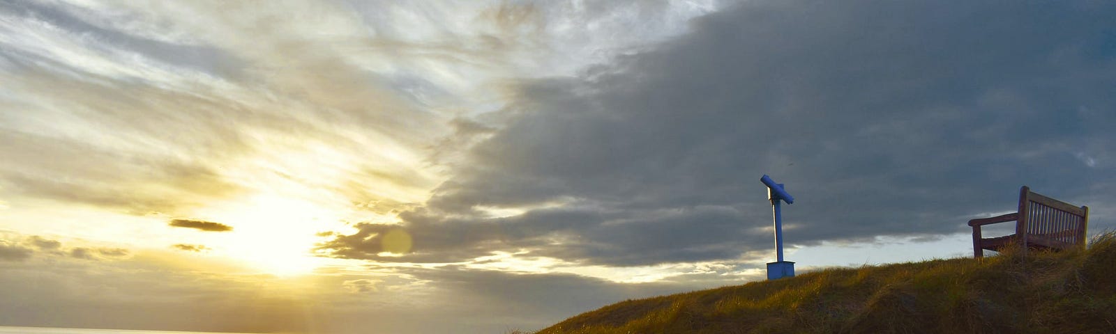 Colour photograph of the green grass at Aberystwyth castle at sunset with a blue telescope and wooden bench in theforeground