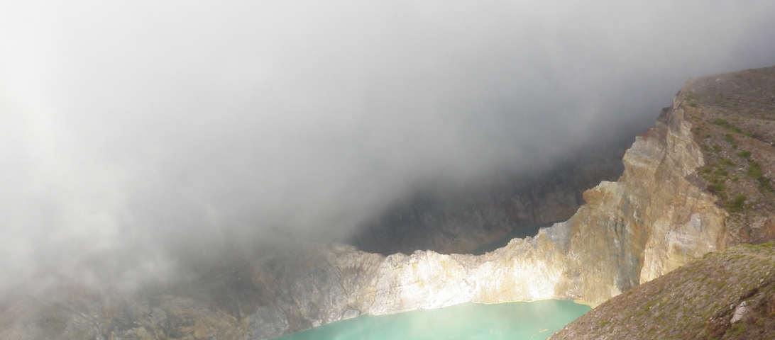 Brown and white cliffs surround a small, pale green lake. A foggy cloud moves diagonally across the photo