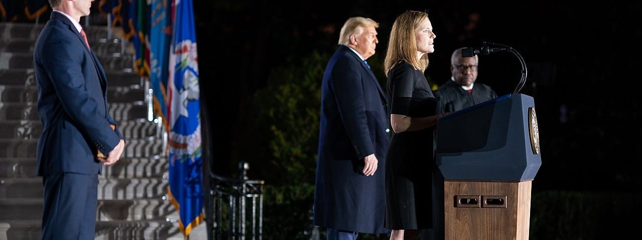 Amy Coney Barrett stands before a podium and microphone, speaking at her swearing-in ceremony. President Donald Trump and Supreme Court Judge Clarence Thomas are on stage with her.