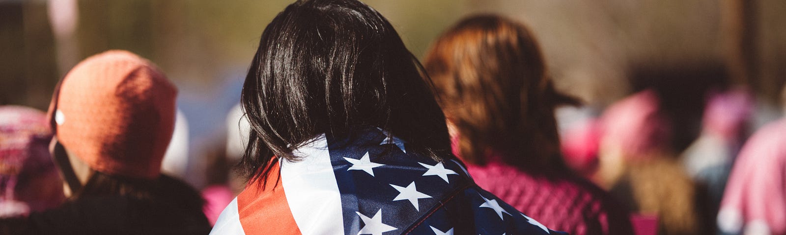 Dark haired woman with back to the camera marches at protest, wrapped in the American flag.