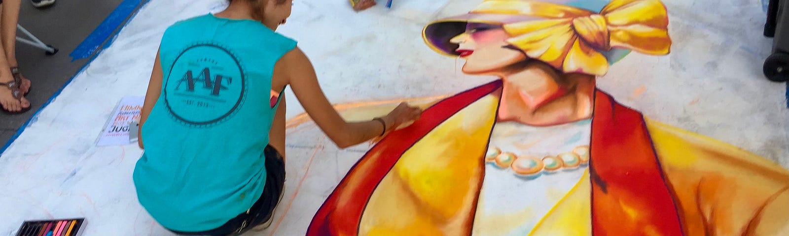 A young dark-haired woman squats down to do more work on a chalk drawing of a 1920s woman in a yellow and red outfit.