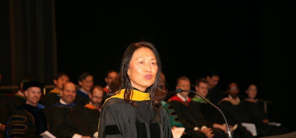Woman dressed in academic regalia speaking at lectern with two seated rows of similarly dressed professors behind her.