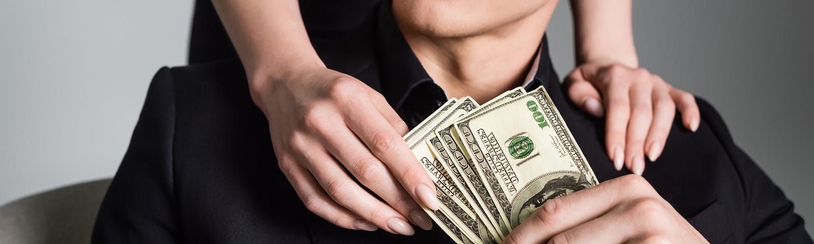 A man in a black suit sitting on a couch holding money as a woman leans over and grabs some of the dollar bills for transactional sex
