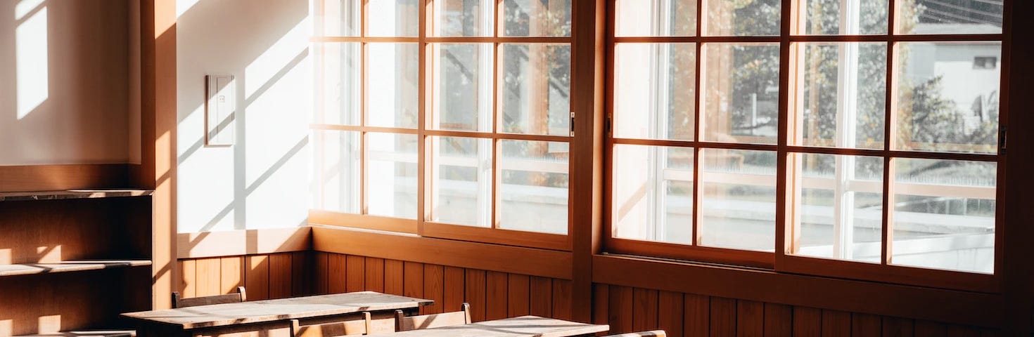A vintage classroom with empty wooden desks and a large window with sunlight shining through.