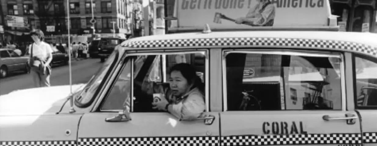 Chinese American woman taxi driver drinking coffee out of the side of a taxi that says “get it done america” on a sign