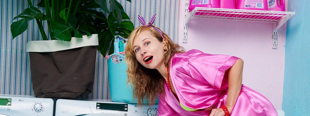 Laundry, woman in pink dressing gown with lingerie underneath bends to take clothing out of washing machine, pink detergents, large pot plant, laundry room, model appears playful or surprised