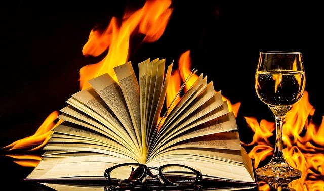 Image of an open book surrounded by flames. In the foreground is a pair of glasses and a drink