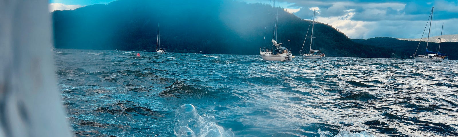 Waves crash into a porthole on a boat. The view is to the ocean and a hill beyond