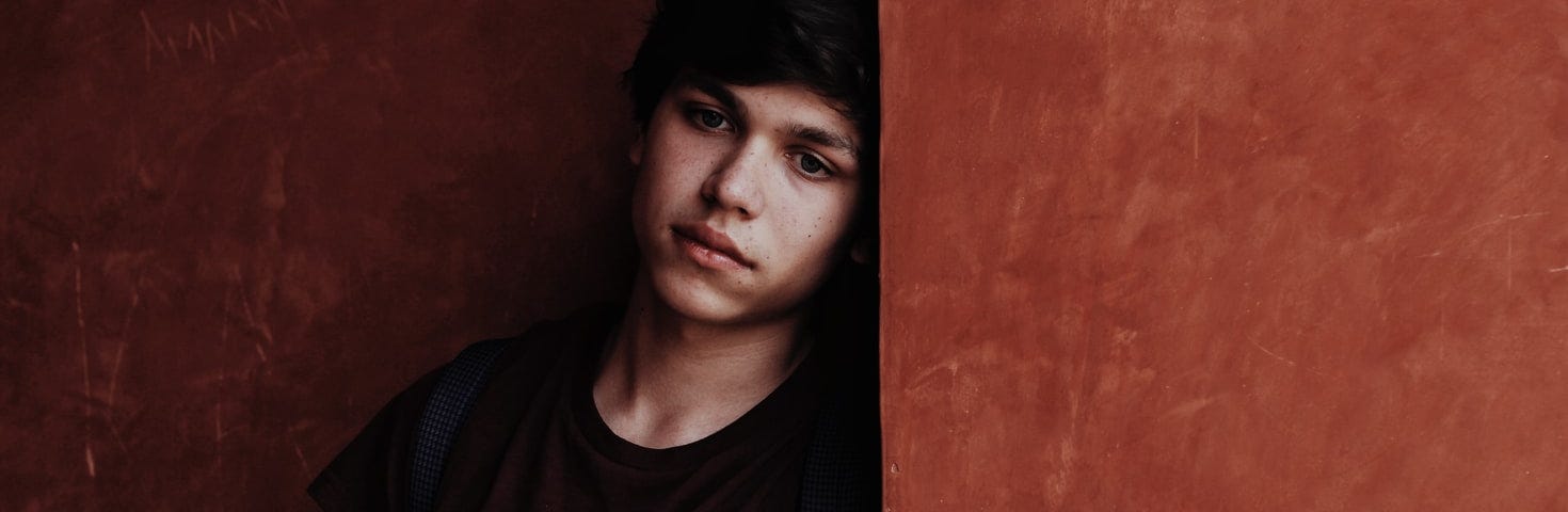 Teenaged boy stands leaning against a wall, looking off into the distance.
