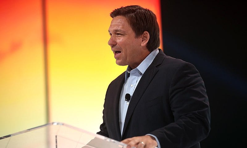 Governor Ron DeSantis speaking with attendees at the 2021 Student Action Summit hosted by Turning Point USA at the Tampa Convention Center in Tampa, Florida. DeSantis is standing at a clear podium and appears to be speaking. He is wearing a black suit and is standing in an orange and yellow background.
