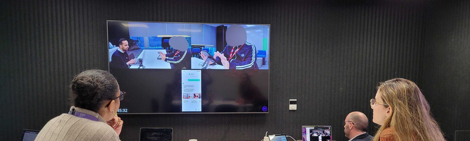 In an office, 3 user researchers are watching a live video feed attached to the wall. On the feed is a split screen, showing 2 users interacting with new digital products and describing their experiences (their faces are blurred) The researchers have pens and post-it’s in front of them as they take notes.