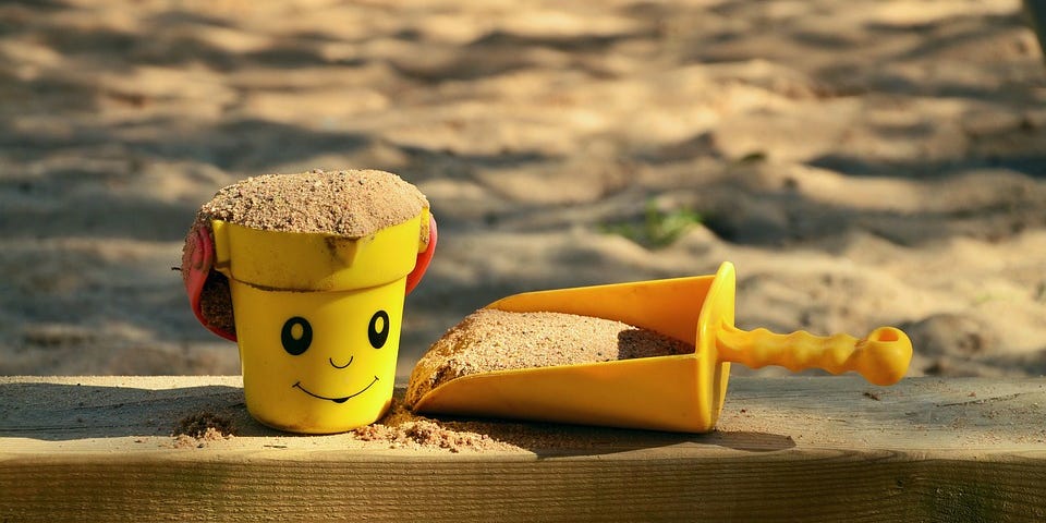 A sand box with a yellow bucket, that shows a smiling face, and a shovel next to it.