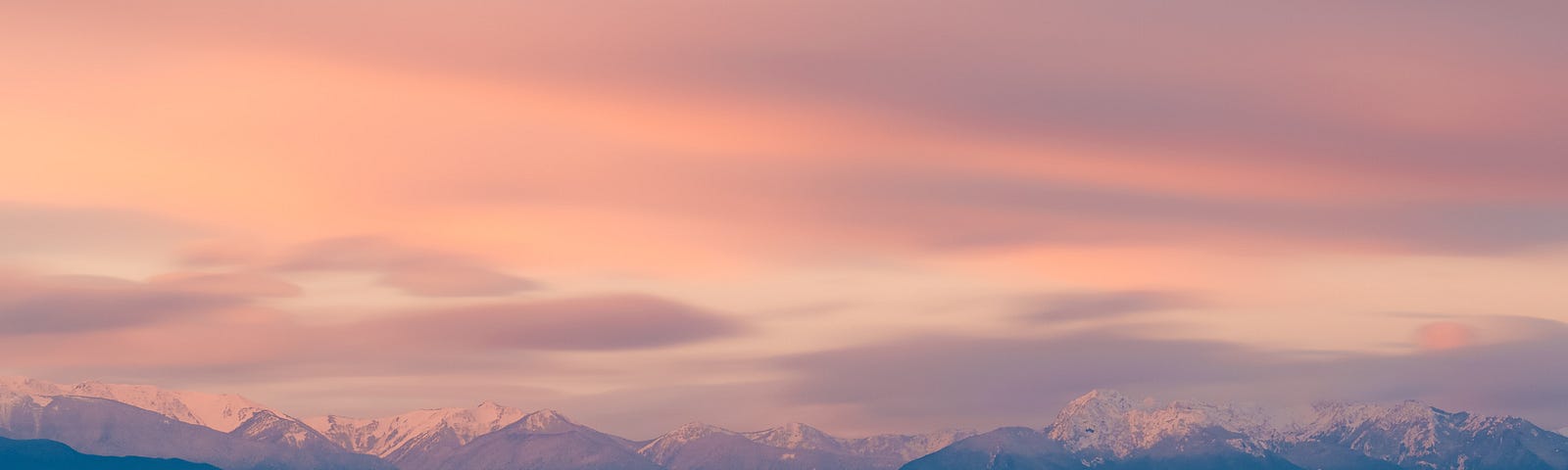 A lake in front of a mountain panorama in the background. The sky is covered in candy-coloured clouds at dusk.