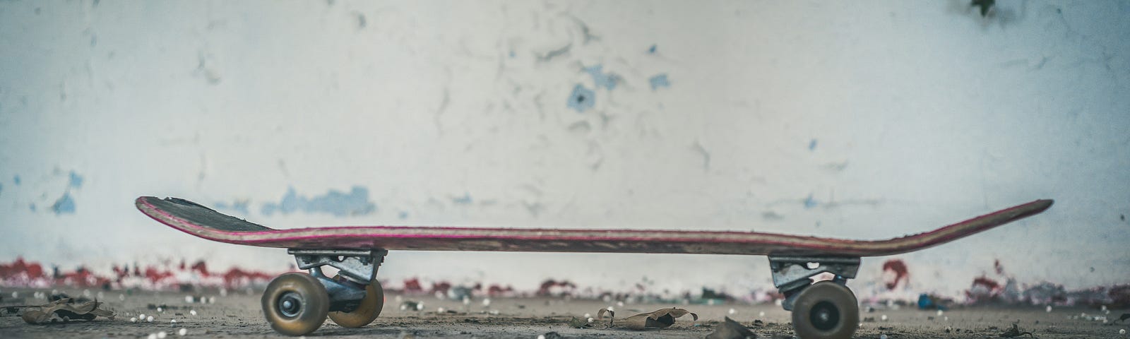 An old-school skateboard sits on the dirt-covered pavement, with a white wall with paint peeling in the background.