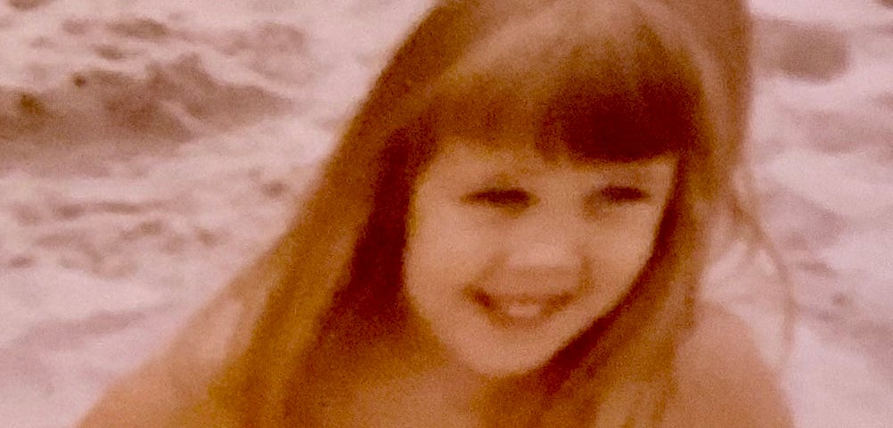 Author Wendi Gordon as a young child sitting in the sand by the sea and smiling.