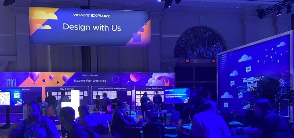 The Design with Us — Illustrate your Enterprise installation in the Explore Hub area was a wide open area with boards on the walls, monitors explaining what was happening and people at tables in a room with purple lighting.