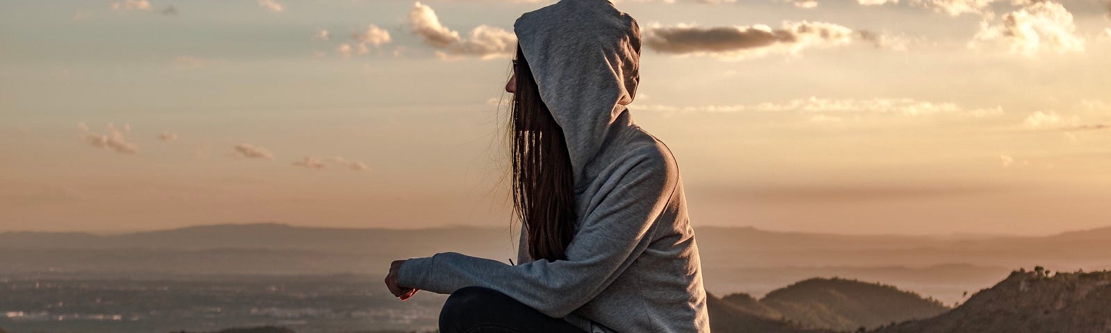 A woman with long dark hair contemplates her life with others as she stares over the edge of a mountain at sunset.