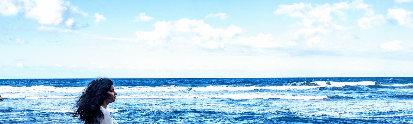 A curly haired woman sits profile with her legs crossed on a dead tree trunk, surrounded by black stones on a black sand beach.