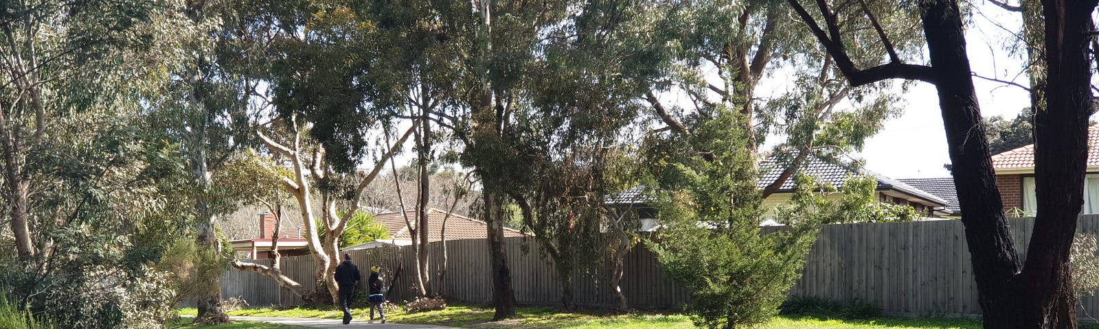 Adult man and young girl walking away from the camera on a footpath through lots of trees and a grassy area. Board privacy fence and houses beyond in the background. No roadmap out of lockdown at the time this photo was taken. Photo by Ann Leach.
