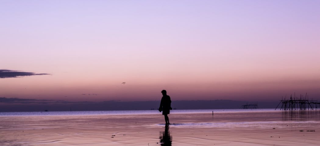 A person walks solemnly on beach, alone, at twilight.