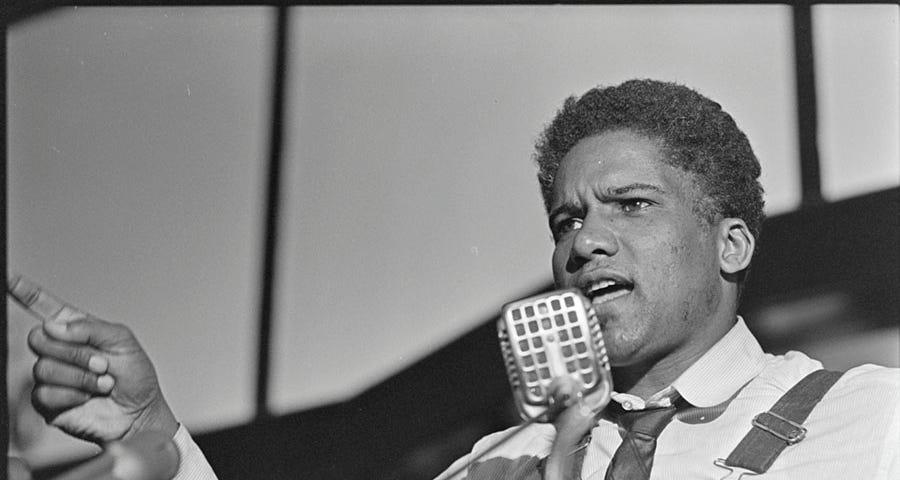 James Forman, a Black man, is dressed in a dress shirt and tie with overalls. He is speaking into a microphone and making a gesture with his right hand, index finger pointed out.