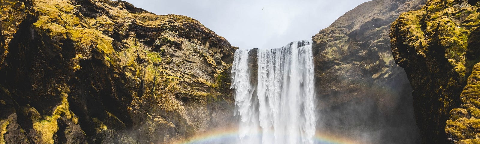 Stream of water falling downward from a cliff creating a waterfall with a rainbow behind it