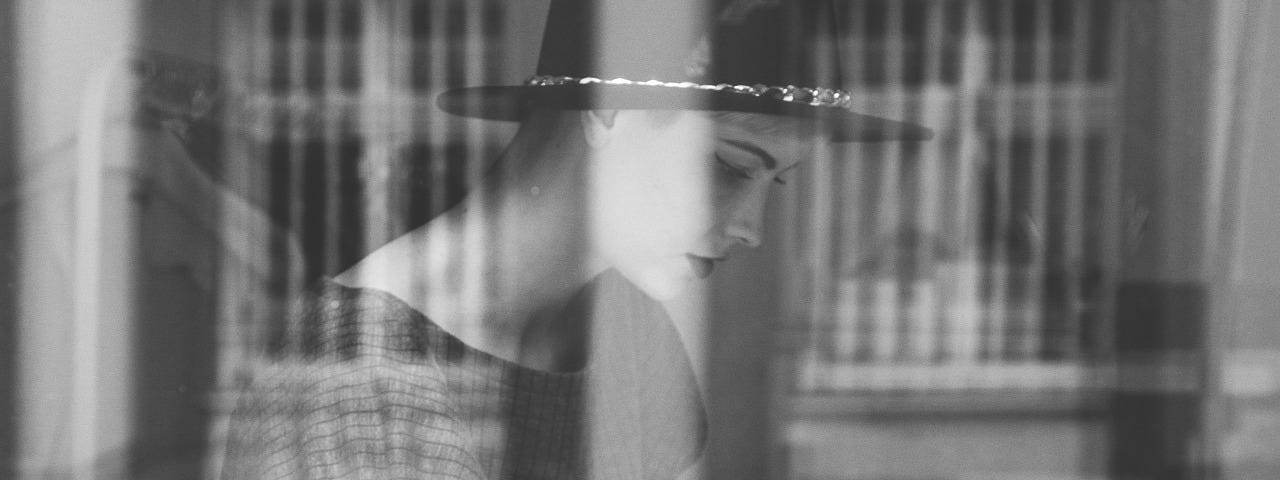 Young woman, stylish, wears hat and has tattoos, reading magazine, surrounded by other magazines, viewed through a window pane which reflects some image.