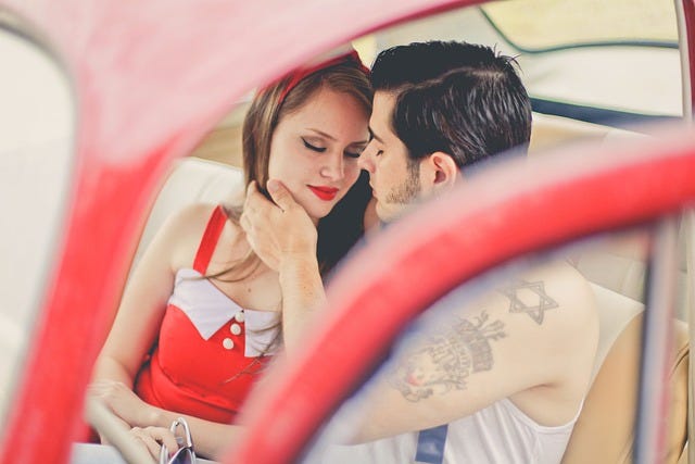 Couple, young man and woman, sitting inside a red car, tattoos, vintage style of dress, impending kiss