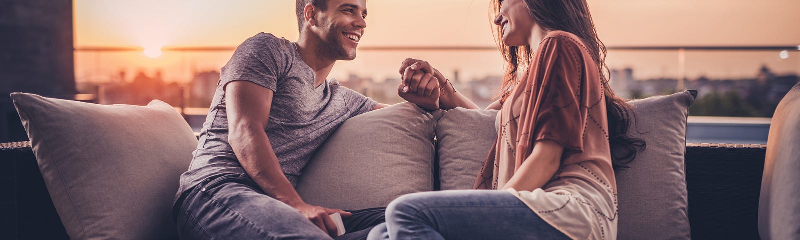 A man and a woman sit together on a roof terrance while the sun is setting. They are holding hands and smiling at each other, but it feels romantic rather than sexual.