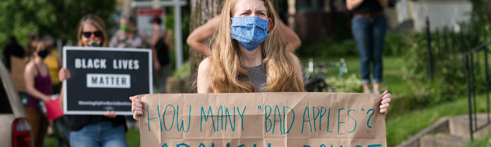 A masked woman holds a sign reading “HOW MANY ‘BAD APPLES’? ABOLISH THE POLICE”. In the background, another woman holds a sign reading “BLACK LIVES MATTER”.