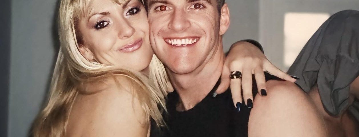 A young, happy couple smiles at the camera. The woman has long blonde hair, and the man’s short brown hair is buzz-cut. They both wear black singlet tops.
