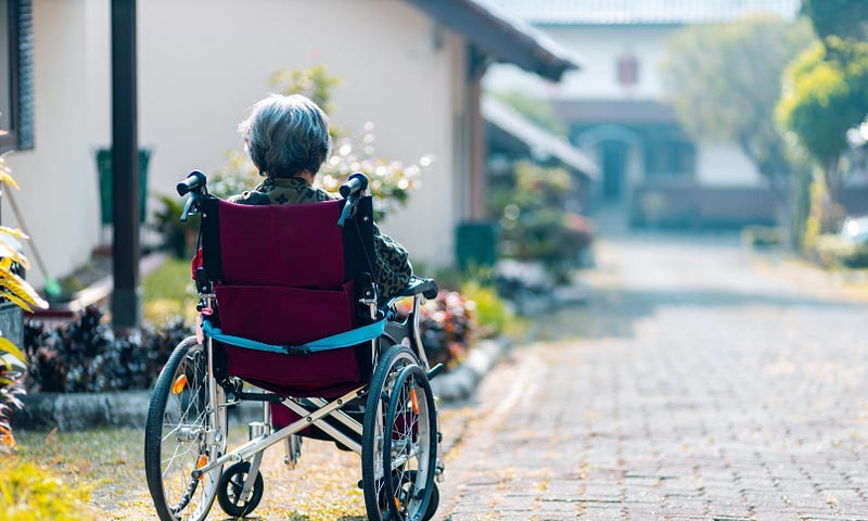 An elderly woman in a wheelchair sitting alone outside of a nursing home in the courtyard