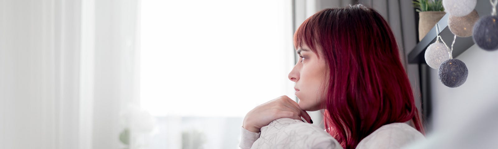 A pensive woman with bright red hair stares out of the window and hugs a pillow.