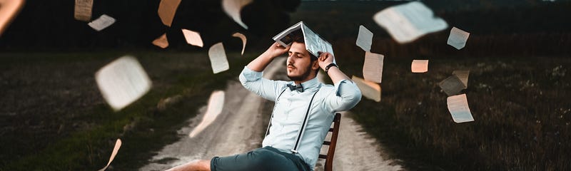 A man sitting on a chair in middle of a road while holding a book on his head and papers all around him
