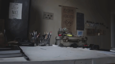 Mostra uma mesa de trabalho organizada com diversos materiais de desenho, incluindo potes com lápis e canetas, um caderno aberto e uma prancheta. Ao fundo, há uma parede decorada com pôsteres inspiradores, um dos quais diz “explore, create, repeat.” A animação mostra uma figura entrando em cena e pulando para fora da mesa, adicionando um elemento divertido e inesperado ao ambiente de trabalho criativo.