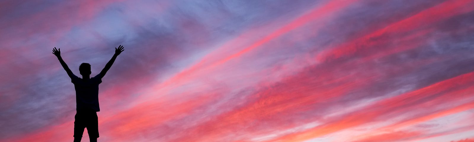 Man standing on a rocky area with arms raised in triumph, silhouetted by a colorful sky at dusk.