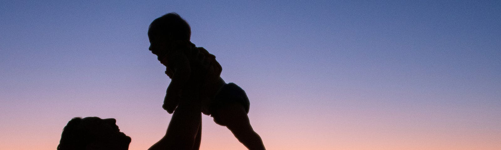 Color photo. In the foreground, a father lifts his baby son up above his head. In the background is a stunning sunset.
