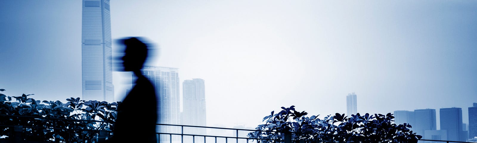 A blurry image of a man walking across an outdoor balcony space. There are tall buildings in the background.