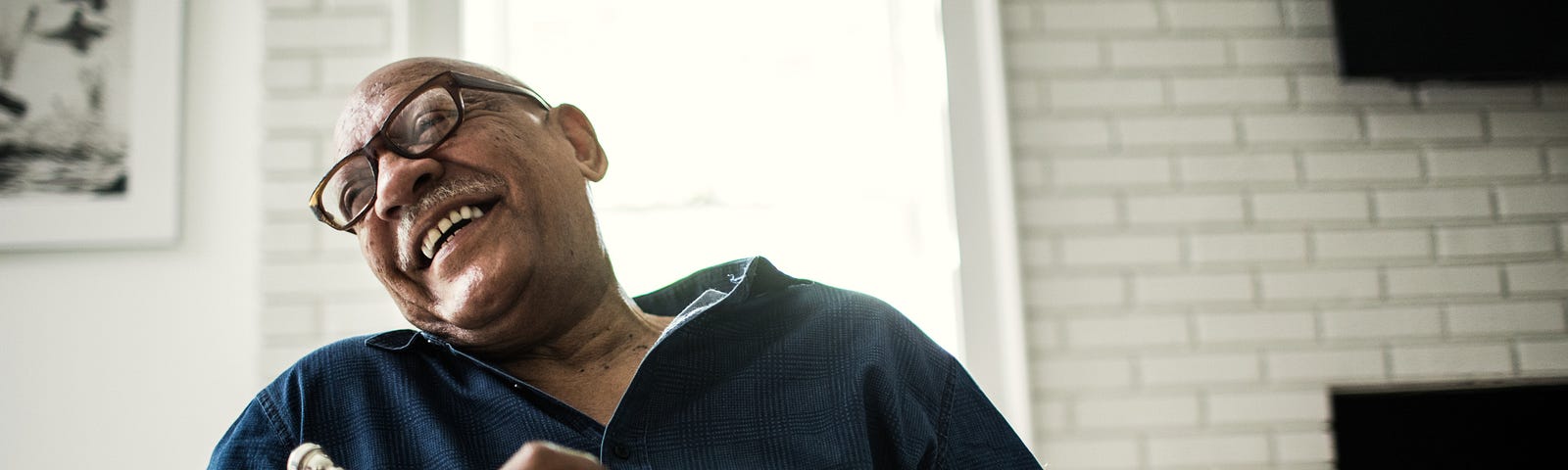 A photo of an older black man holding a trumpet in his living room. He is laughing.