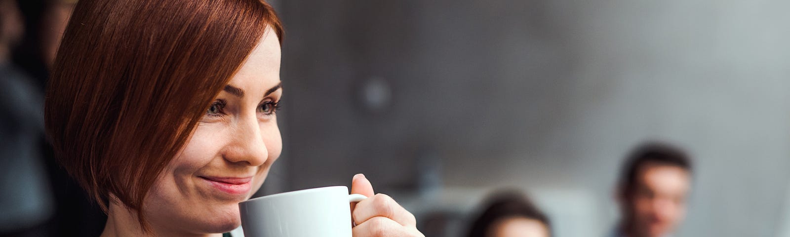 A woman smirks as she holds a coffee mug up to her face in front of 2 coworkers.