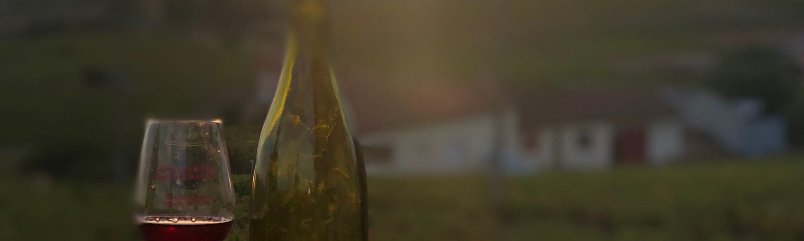 A bottle and glass of red wine with vineyards and sunset in the background