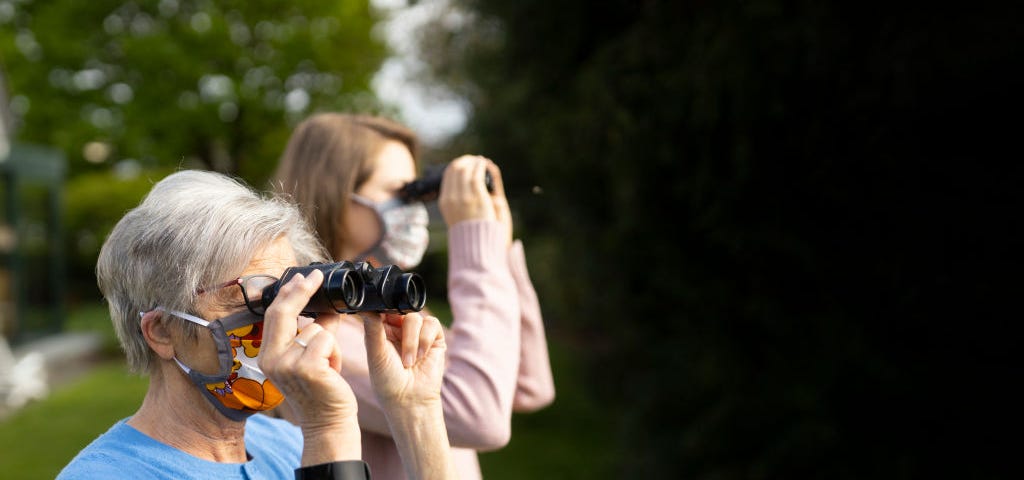 Two women wearing face masks use binoculars to look in the distance.