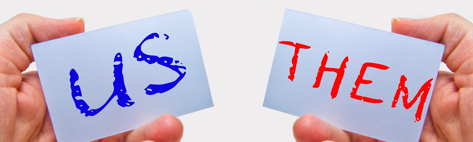 An image of two hands holding signs reading “us” in blue and “them” in red. “Us” is on the left. “Them” are on the right.