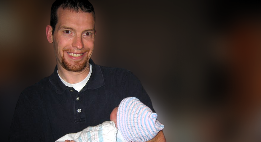 Father holds swaddled baby shortly after birth with mother in the background