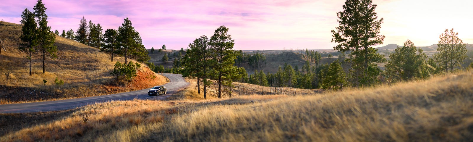 My Mazda sits on a small section of pavement between the trees and grasses of the Black Hills as the sun sets just out of camera’s view.