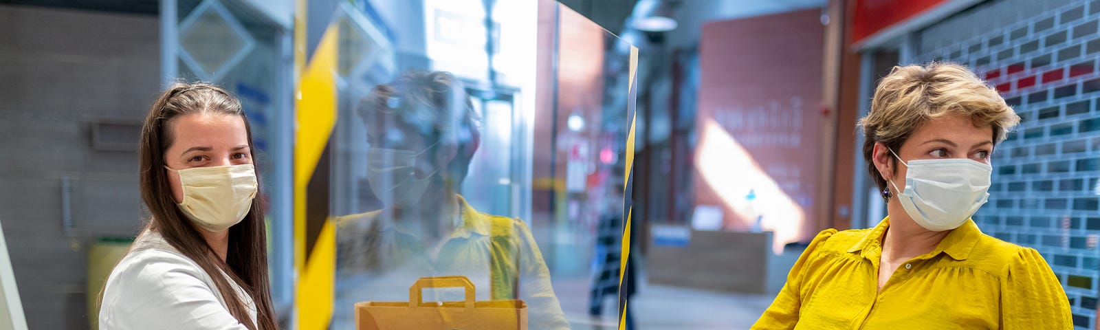 Woman wearing a face mask and gloves gives her credit card to a cashier on the other side of a plexiglass at checkout.