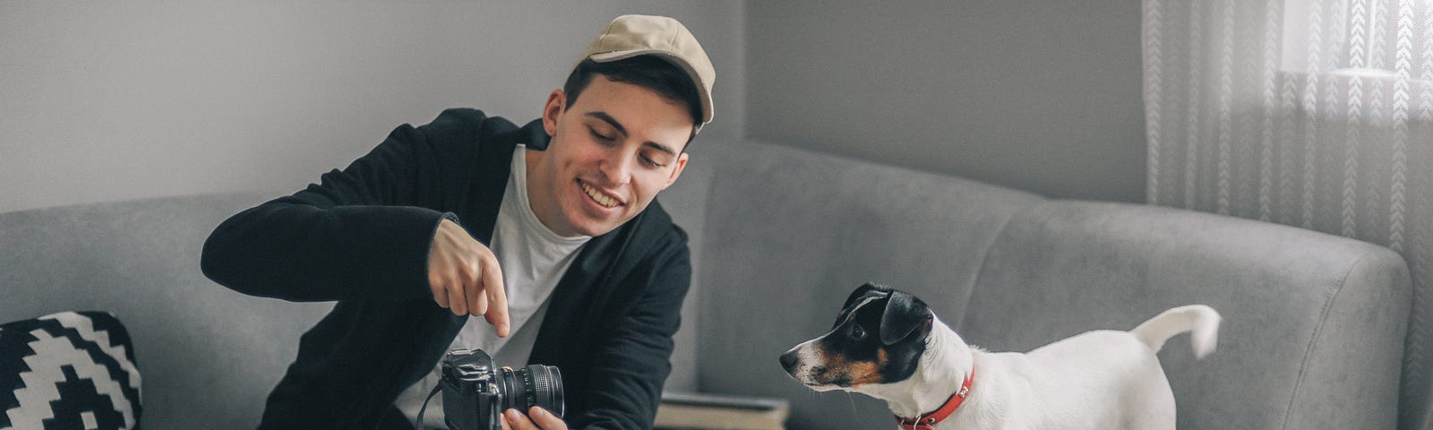 A young man sets up his camera and tripod on his couch. His dog is on the couch looking at him hold his camera.